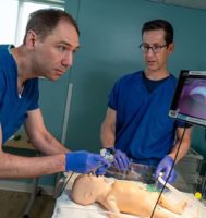 A doctor wearing blue scrubs works on a pediatric manikin patient while another doctor looks on.