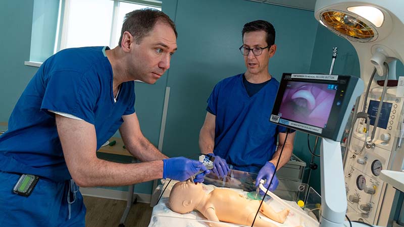 A doctor wearing blue scrubs works on a pediatric manikin patient while another doctor looks on.