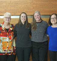 A group of six women standing against a wood wall with names on it.