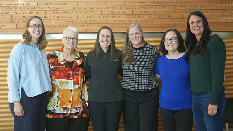 A group of six women standing against a wood wall with names on it.
