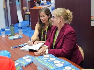 Two women sitting at a table and signing copies of books.